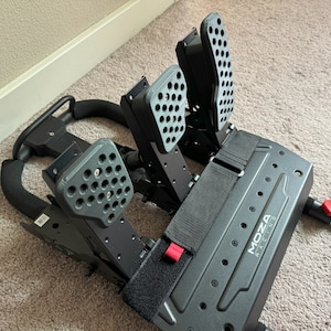 May include: Black racing pedals with a Moza Racing logo. The pedals have a textured surface and are attached to a black metal frame.