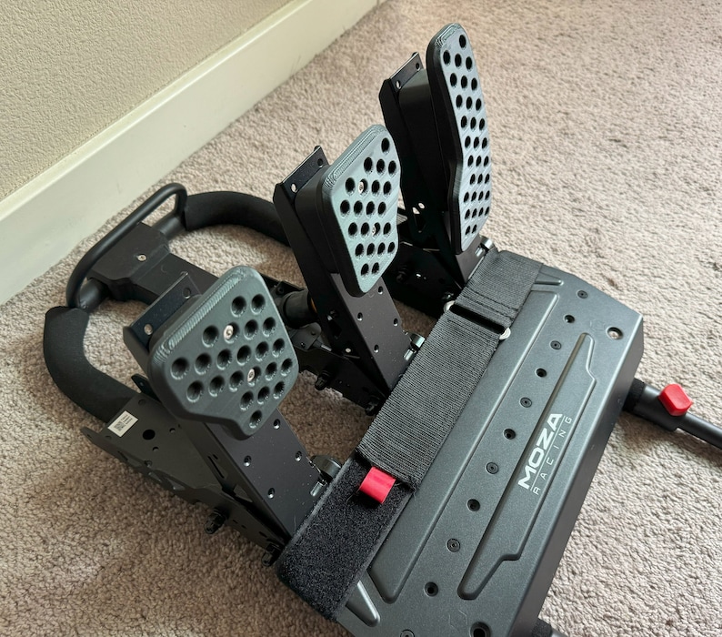 May include: Black racing pedals with a Moza Racing logo. The pedals have a textured surface and are attached to a black metal frame.