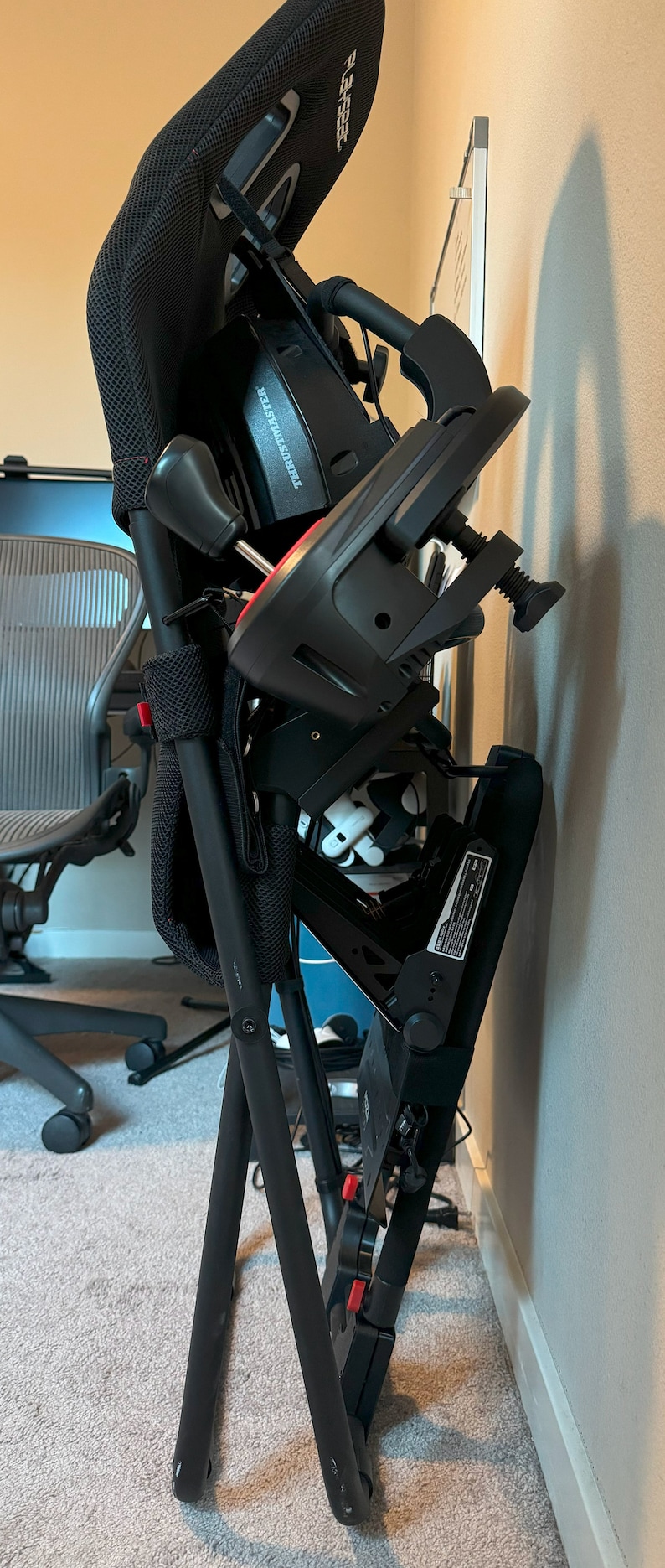 K&ouml;nnte beinhalten: Ein schwarzer Rennsitz-Simulator mit einem schwarzen und roten Lenkrad und Schaltknauf. Der Stuhl ist zusammengeklappt und an einer Wand angelehnt.