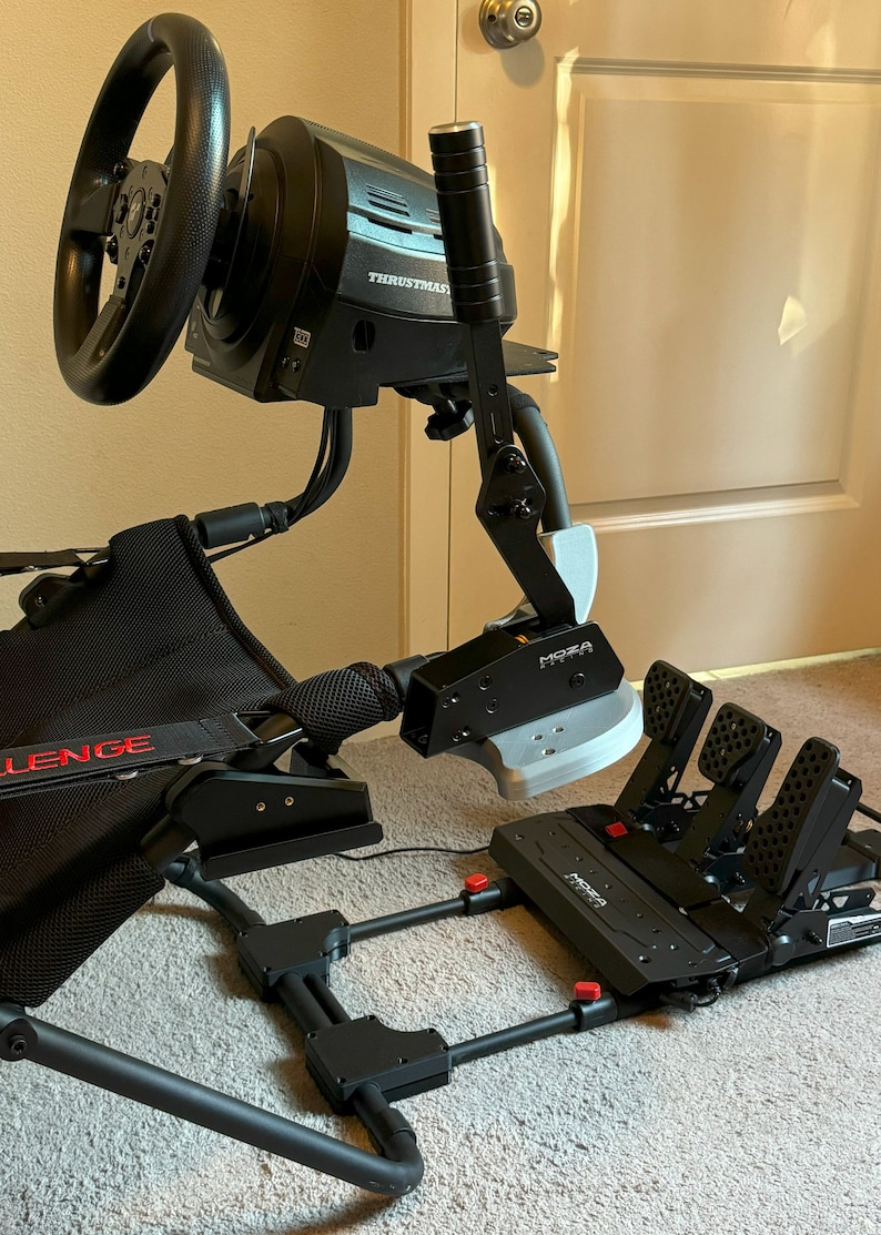 K&ouml;nnte beinhalten: Ein schwarzes Rennlenkrad- und Pedalset mit einem Schalthebel. Das Lenkrad hat einen schwarzen Ledergriff und die Pedale haben einen schwarzen Metallrahmen mit roten Kn&ouml;pfen. Das Set ist auf einem schwarzen Metallrahmen mit einem schwarzen Netz-Sitz montiert. Der Text "THRUSTMASTER" ist auf dem Lenkrad und "MOZA" auf den Pedalen sichtbar.