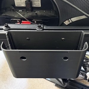 May include: A black, U-shaped plastic mounting bracket with a rectangular base and two circular holes on the front panel. The bracket is attached to a horizontal metal frame rail, featuring a textured surface and visible screw heads for secure installation.