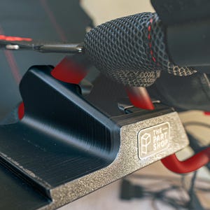 May include: Close-up of a black metal bracket with a red strap attached. The bracket has a textured surface and a logo that reads "The Part Shop".
