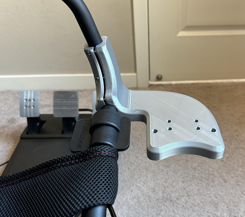 K&ouml;nnte beinhalten: Ein silberfarbener 3D-gedruckter Rennlenkradhalter mit einem schwarzen Gurt und einem schwarzen Netzstoff. Der Halter hat eine gebogene Form und mehrere L&ouml;cher.