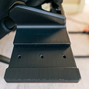 May include: Close-up of a black plastic bracket with four holes. The bracket is attached to a black metal frame.