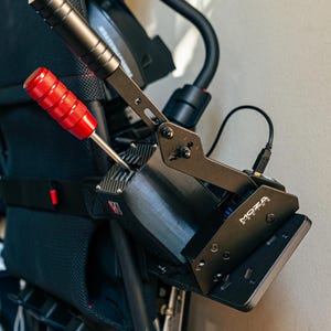 May include: A black and silver racing shifter with a red handle attached to a black metal frame. The frame has the word "MOZA" printed on it.