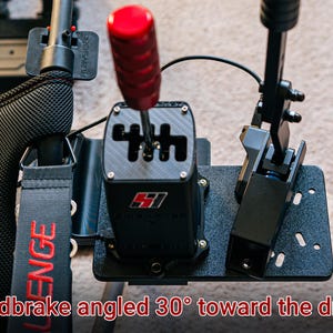May include: A black racing simulator shifter with a red gear knob and a black handbrake angled 30 degrees towards the driver. The shifter has a carbon fiber look and the text "SH1FT" is printed on the front.