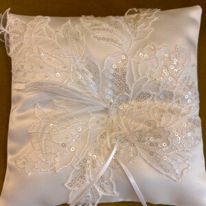 May include: White satin ring pillow with a delicate lace design and sequins. The pillow has a white satin ribbon tied around it.