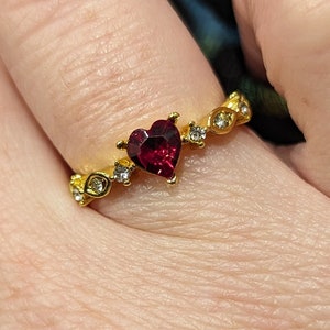 May include: Gold ring with a red heart-shaped gemstone and smaller clear gemstones on the band.