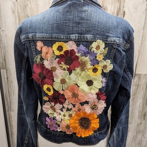 May include: A dark blue denim jacket with a large embroidered floral design on the back. The flowers are in shades of red, orange, yellow, pink, and purple.
