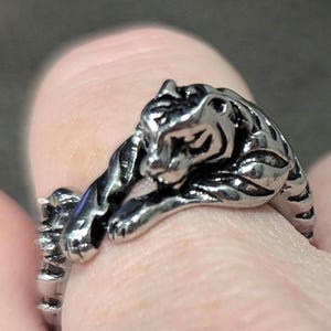 May include: A silver ring featuring a detailed tiger design. The tiger is depicted lying down with its head resting on its paws.