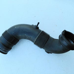 May include: A black automotive air intake hose featuring a curved, flexible rubber construction with ribbed sections for bending, a small protruding nozzle on the top, and circular connection ports at both ends for engine installation.