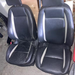 May include: Two black leather car seats with white accent stripes. The seats have headrests and show signs of wear. The seats are likely from a vehicle's interior and are placed on a concrete floor.