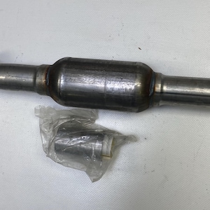 Puede incluir: Un convertidor catalítico metálico con un cuerpo cilíndrico y un tubo de entrada curvado. Un componente más pequeño y envuelto descansa debajo. Las piezas son plateadas y parecen nuevas.