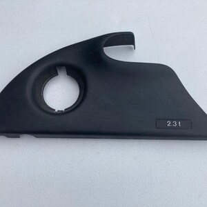 Peut inclure: Un cache-moteur automobile en plastique noir présentant une ouverture circulaire sur le côté gauche et une indentation rectangulaire sur le côté droit avec le texte 2.3 t imprimé en blanc. La pièce a un bord supérieur incurvé et une base plate.