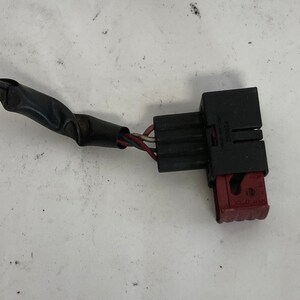 May include: A black electrical connector with red and blue wires. The connector has a black plastic housing and a red plastic release lever. The wires are wrapped in black electrical tape.