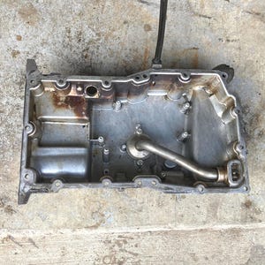 Op de afbeelding: Een gebruikte, zilverkleurige motoroliepan met een gebogen pijp en verschillende boutgaten. De pan heeft wat roestkleurige vlekken en is rechthoekig met afgeronde hoeken. Een zwarte slang loopt van bovenaf.