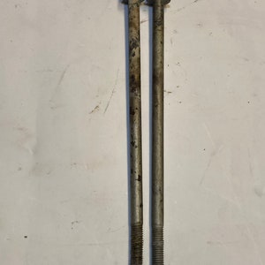May include: Two long, metal bolts with hexagonal heads and threaded ends. The bolts are silver with some signs of wear and rust. They are positioned vertically against a white background, showcasing their length and design.