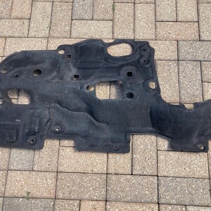 May include: A black, irregularly shaped automotive part, possibly a sound deadening panel or undercarriage component. It features multiple holes and mounting points, and is made of a textured, possibly felt-like material. The part is lying on a brick surface.