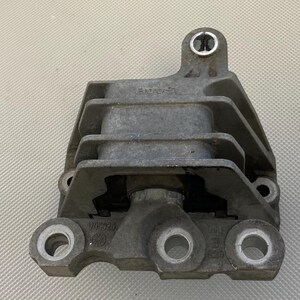 May include: A gray metal engine mount with multiple bolts and a rubber insert. The mount has a circular hole on the top and a rectangular hole on the bottom.
