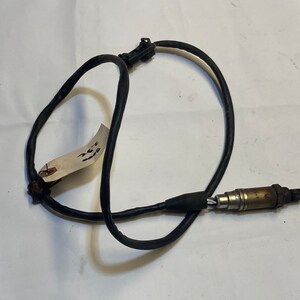 Może przedstawiać: Czarny czujnik tlenu z drutem, srebrną końcówką i czarnym złączem. Czujnik jest zwinięty i ma białą etykietę z napisem "261".