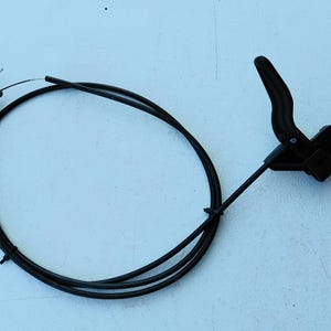 May include: A black mechanical control cable assembly featuring a curved plastic lever handle attached to a long, flexible housing. The cable ends are fitted with small metal cylindrical stoppers and exposed wire tips for connection.