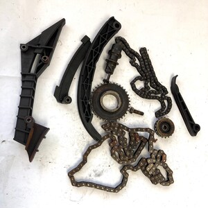 以下が含まれることがあります： 黒と金属製のエンジン部品の詰め合わせ。チェーン、ギア、テンショナーなど、白い背景に配置されています。コンポーネントはサイズと形状が異なり、機械的または自動車的なテーマを示唆しています。
