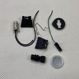May include: Assortment of black plastic car parts, including a connector, a sensor, a switch, and a small round piece with a hole in the center. There is also a silver metal part with a black wire attached.