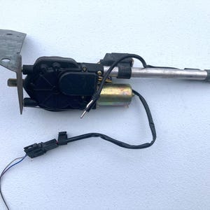 May include: An automotive power antenna assembly featuring a black plastic motor housing, a metallic cylindrical motor, and a long silver telescopic mast. A galvanized steel mounting bracket is attached to the side, with black wiring and a connector.