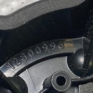 May include: Close-up of a black plastic component with the number "12800996" embossed on its surface. The component has a circular hole and a rounded, protruding feature. The image is well-lit, highlighting the details of the molded plastic.