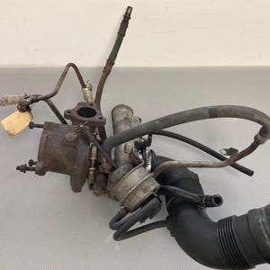 May include: A close-up of a used car part, a turbocharger with a black rubber hose and metal pipes. The turbocharger is rusty and has a label attached to it.