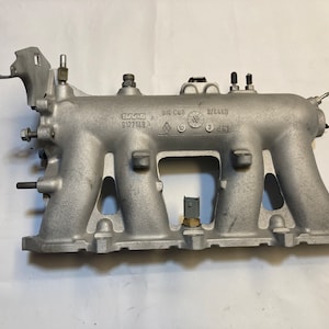 May include: A silver aluminum intake manifold with a black plastic sensor and the text "SAAB 9172148" and "Sie Cu3 8F6649" printed on it.