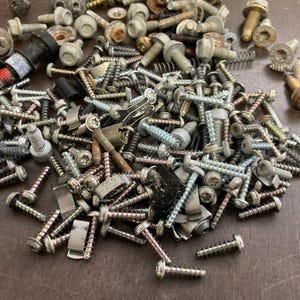 May include: A large pile of assorted screws, bolts, and other metal fasteners in various sizes and finishes. The fasteners are scattered on a brown surface.