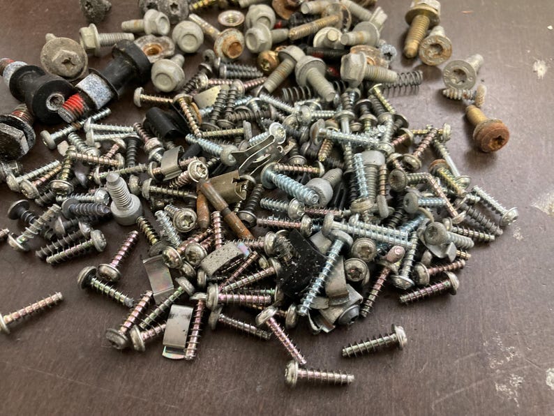 May include: A large pile of assorted screws, bolts, and other metal fasteners in various sizes and finishes. The fasteners are scattered on a brown surface.