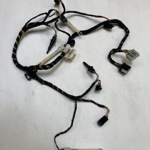 May include: A black and multi-coloured automotive wiring harness with various connectors and sensors. The harness includes multiple wires, connectors, and sensors, with a few white plastic components. The image is taken from a top-down perspective.