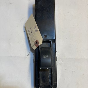 Puede incluir: Interruptor de ventana de coche de plástico negro con un solo botón y un icono de ventana. El interruptor está unido a una carcasa de plástico negro.