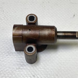 May include: A brown metal mechanical part with a cylindrical body, two cylindrical extensions, and a rectangular end. The part has a worn, aged appearance, suggesting use. The object is likely a component from a larger machine or device.
