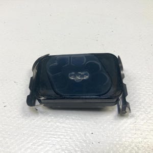 May include: A black rectangular object with rounded corners and a textured surface. The top features a design with three connected circles. Two metal brackets are attached to the sides.