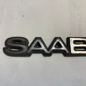 May include: A black plastic Saab emblem with silver lettering. The letters are raised and have a textured finish.