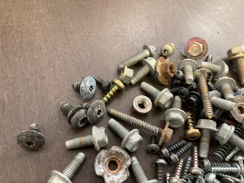 May include: A close-up of a variety of screws and bolts, some with a silver finish and others with a gold finish. The screws and bolts are scattered on a brown wooden surface.
