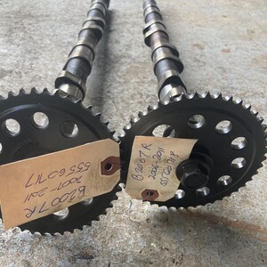 May include: Two automotive camshafts with attached sprockets. The sprockets are black with multiple holes and a toothed outer edge. Each camshaft has a series of lobes. Paper tags are attached to the sprockets with handwritten text.