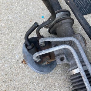 May include: Close-up of a used automotive power steering rack and pinion assembly. The metal components show signs of wear and rust. Various tubes and fittings are attached, with a focus on the hydraulic lines and the steering gear housing.