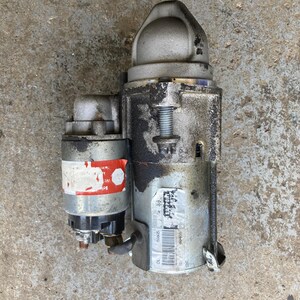May include: A used automotive starter motor, featuring a cylindrical body with a smaller attached component. The metal casing shows signs of wear, with some areas appearing dirty or stained. A label with text is visible on the side.