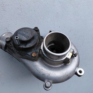 May include: A metallic automotive turbocharger unit featuring a circular housing, a central turbine intake, and a dark circular valve component with a small metal pipe protruding from the top, secured by several visible bolts.