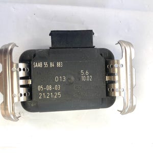 May include: A black electronic sensor with silver metal brackets. The sensor has the text "SAAB 55 84 883", "013", "05-08-03 21:21:25", "5.6 10.02" printed on it.