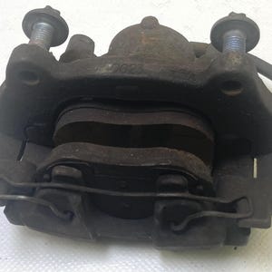 May include: A used, dark-colored brake caliper with visible wear and tear. The caliper has two bolts, a wire clip, and brake pads. The number "2623" is visible on the caliper. This automotive part is likely for a vehicle.