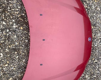 May include: A red car bonnet with a curved design. The bonnet has a smooth, glossy finish and a small emblem on the right side. Three small black fasteners are visible.