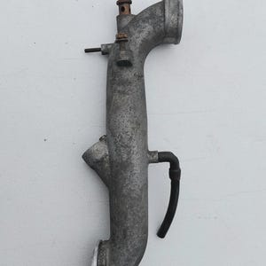 May include: A weathered, gray cast metal automotive intake pipe featuring a curved tubular body, a circular mounting flange at the base, a side-mounted black rubber hose, and several small threaded metal studs and bolts protruding from the top.