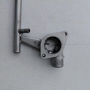 May include: A gray metal automotive coolant pipe assembly featuring a cylindrical tube connected to a flanged housing with a circular opening and a smaller perpendicular outlet port, showing a weathered metallic finish.