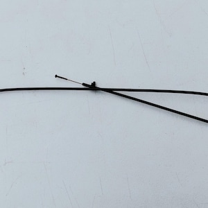 May include: A black automotive control cable featuring a flexible outer housing, a metal mounting bracket at one end, and a thin inner wire cable that extends beyond the housing with a small circular stopper at the tip.
