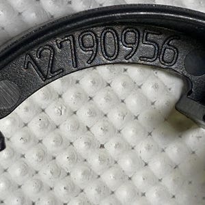 May include: Close-up of a black, curved plastic component with the number "12790956" embossed on its surface. The component has a matte finish and is set against a textured, white background.